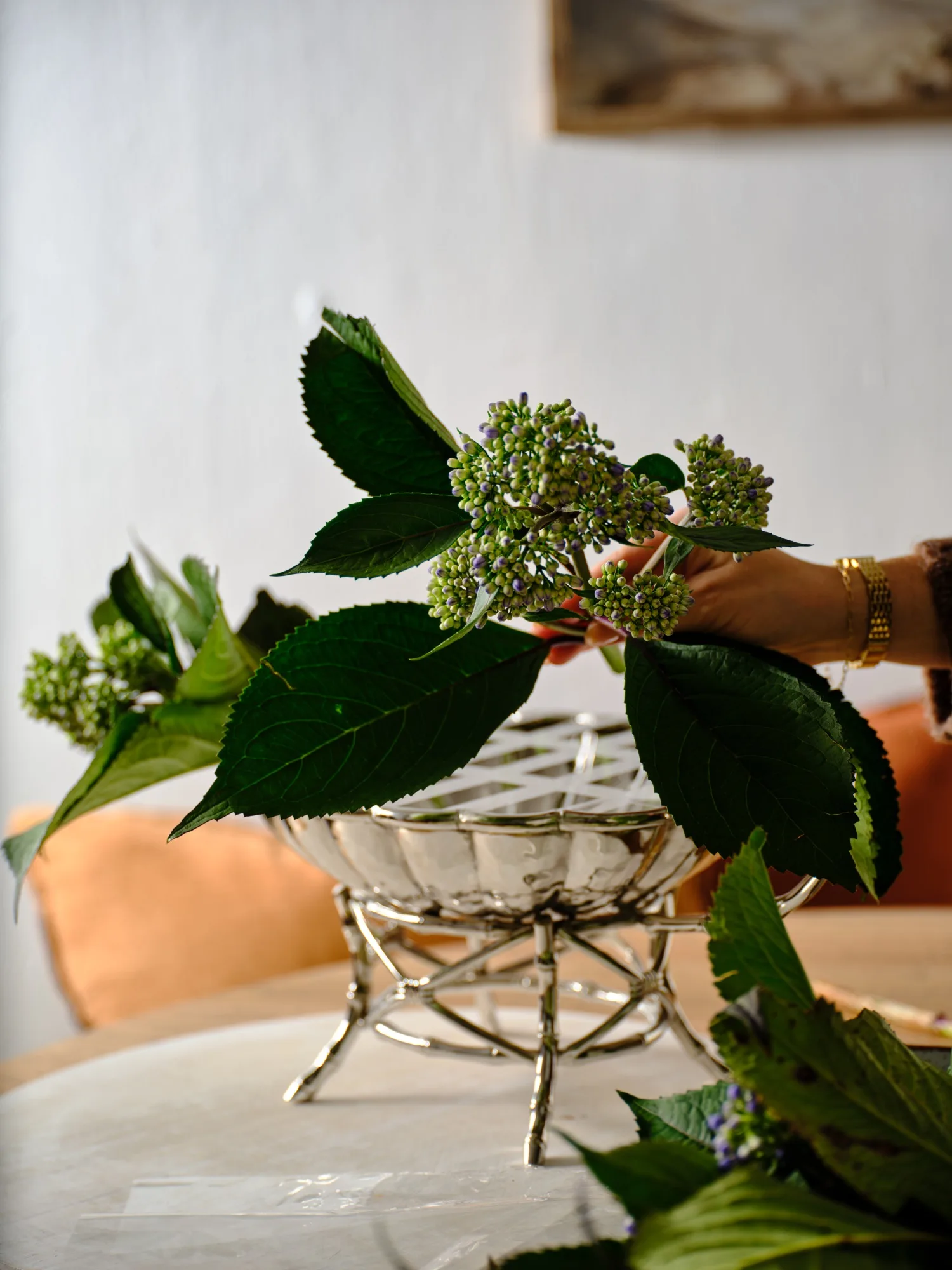 A hand places green hydrangeas in a silver bowl on a table with peach chairs in the background.