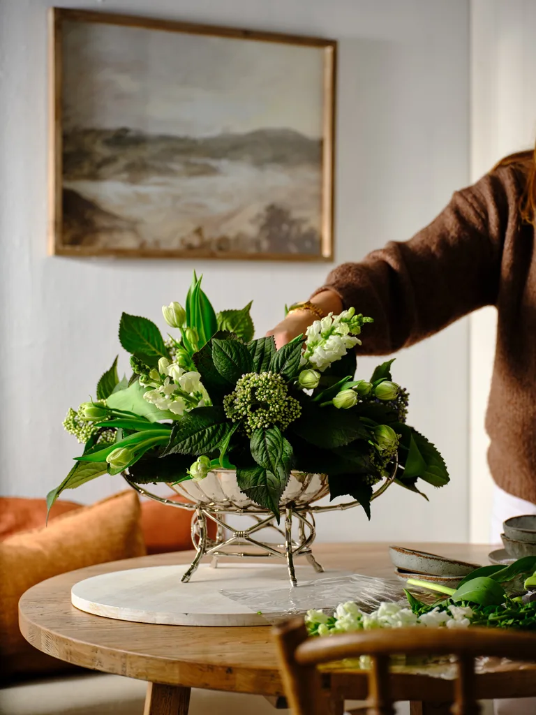 A person arranging a lush green and white floral centerpiece on a wooden table, with a landscape painting in the background.
