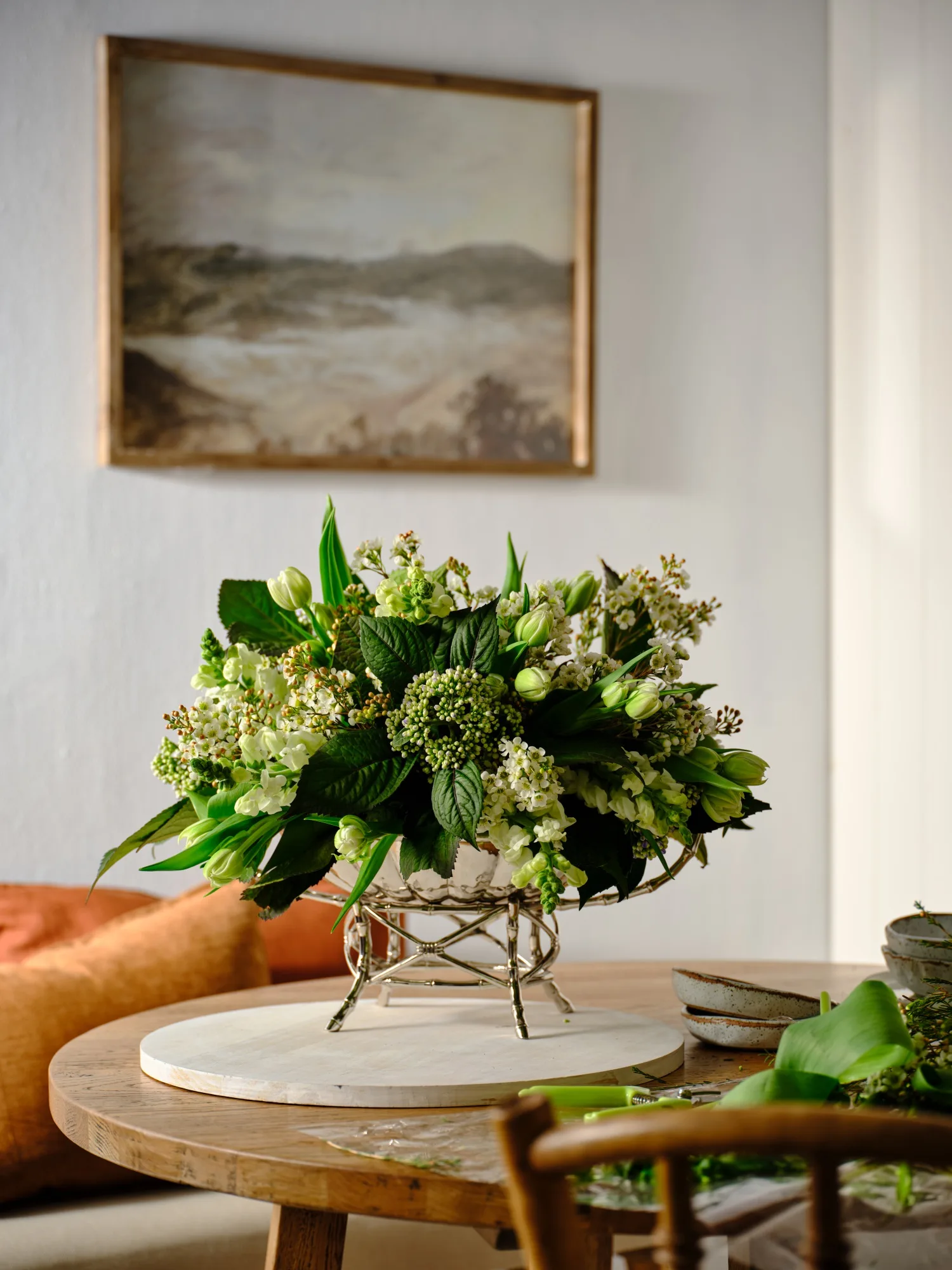 Floral arrangement with green and white flowers on a wooden table, framed landscape painting in the background.