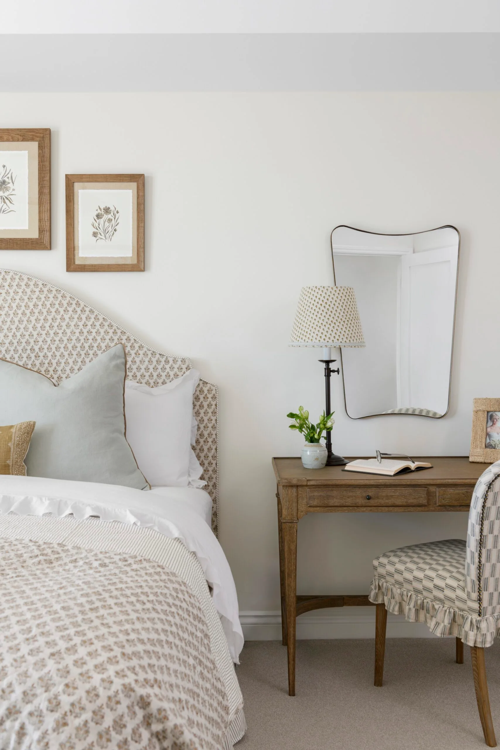 Bedroom with a patterned bedspread, wooden desk with lamp, mirror, and framed art on white walls.