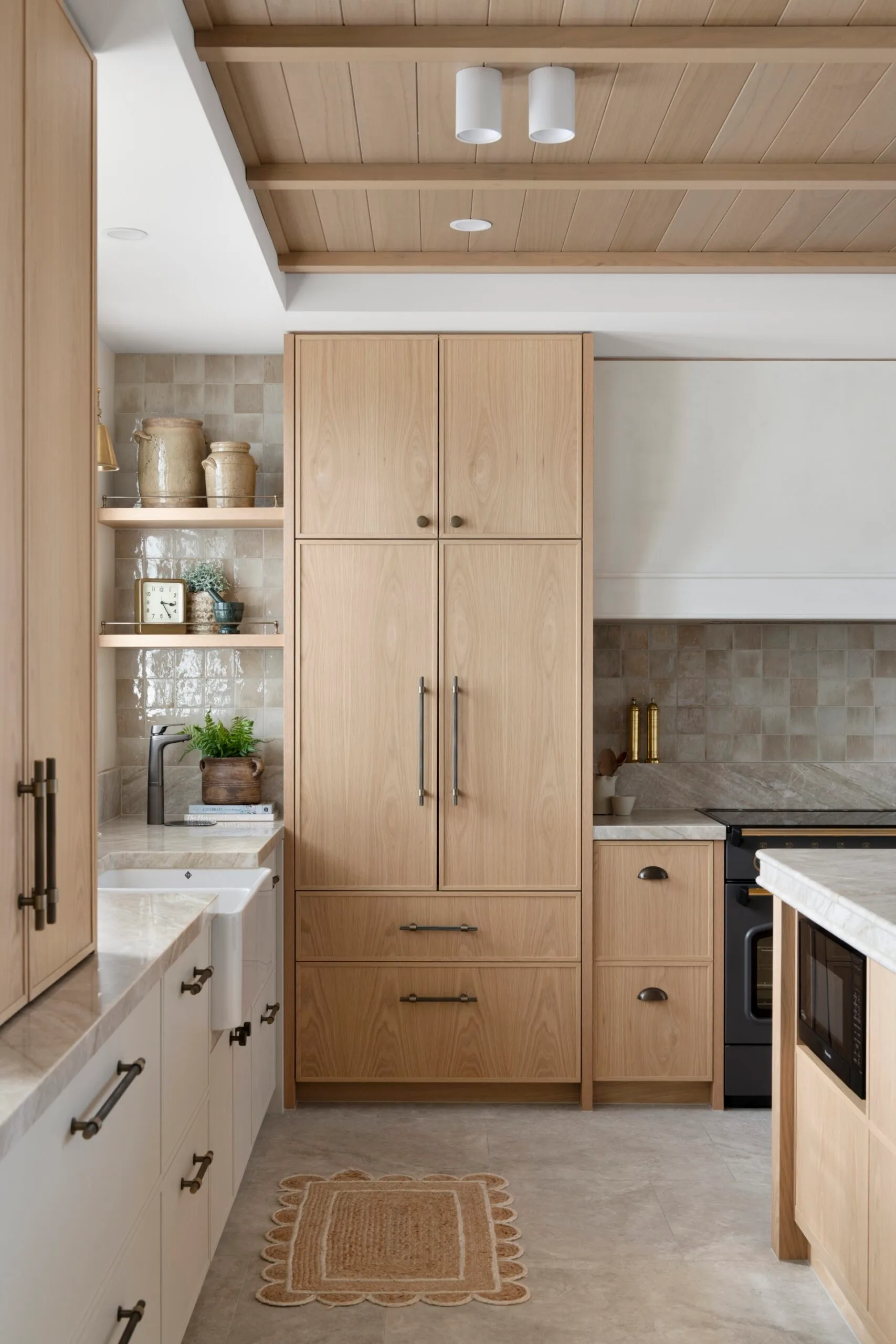 Modern kitchen with light wood cabinets, beige tiles, a black stove, and marble countertops. Rug and decor visible.