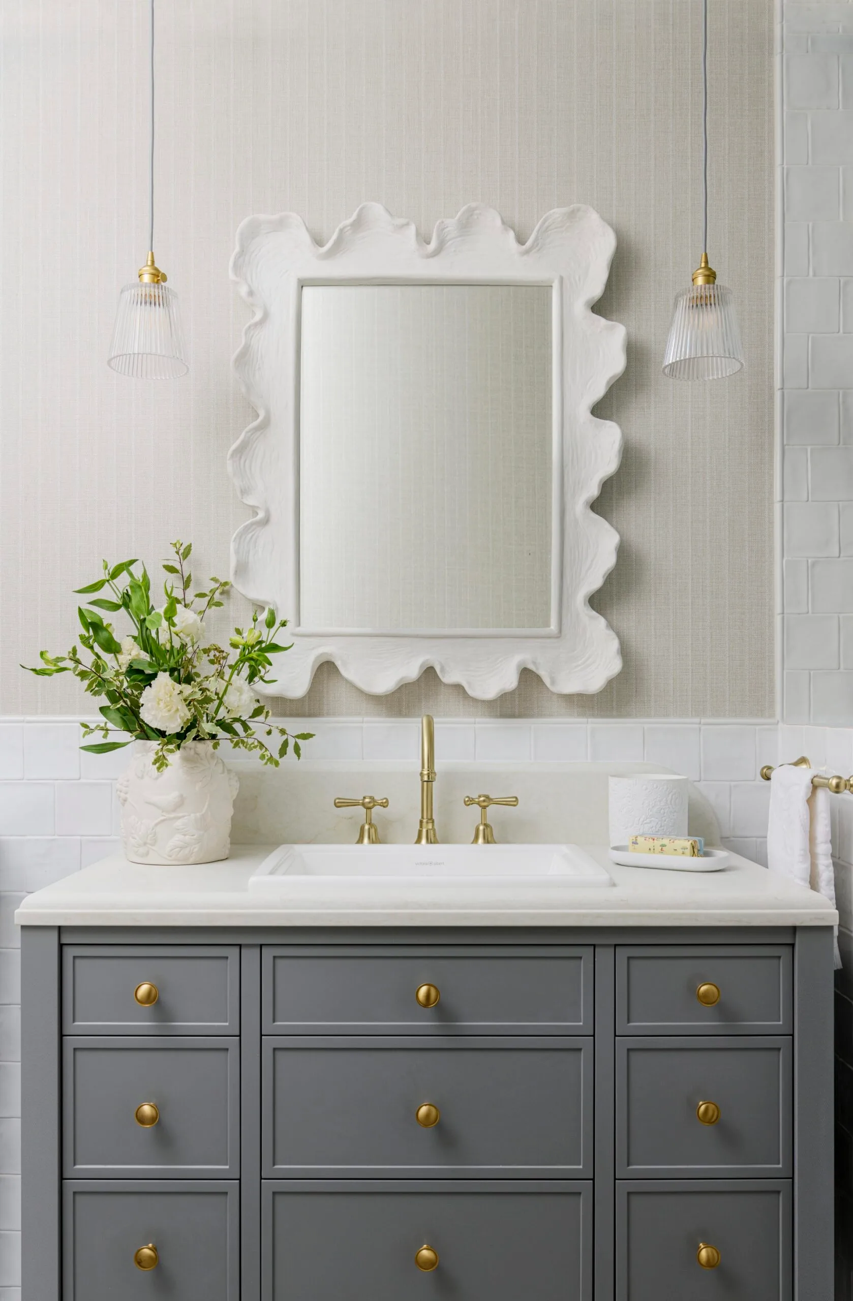 Elegant bathroom vanity with gray cabinets, gold hardware, floral vase, ornate mirror, pendant lights, and white tiles.