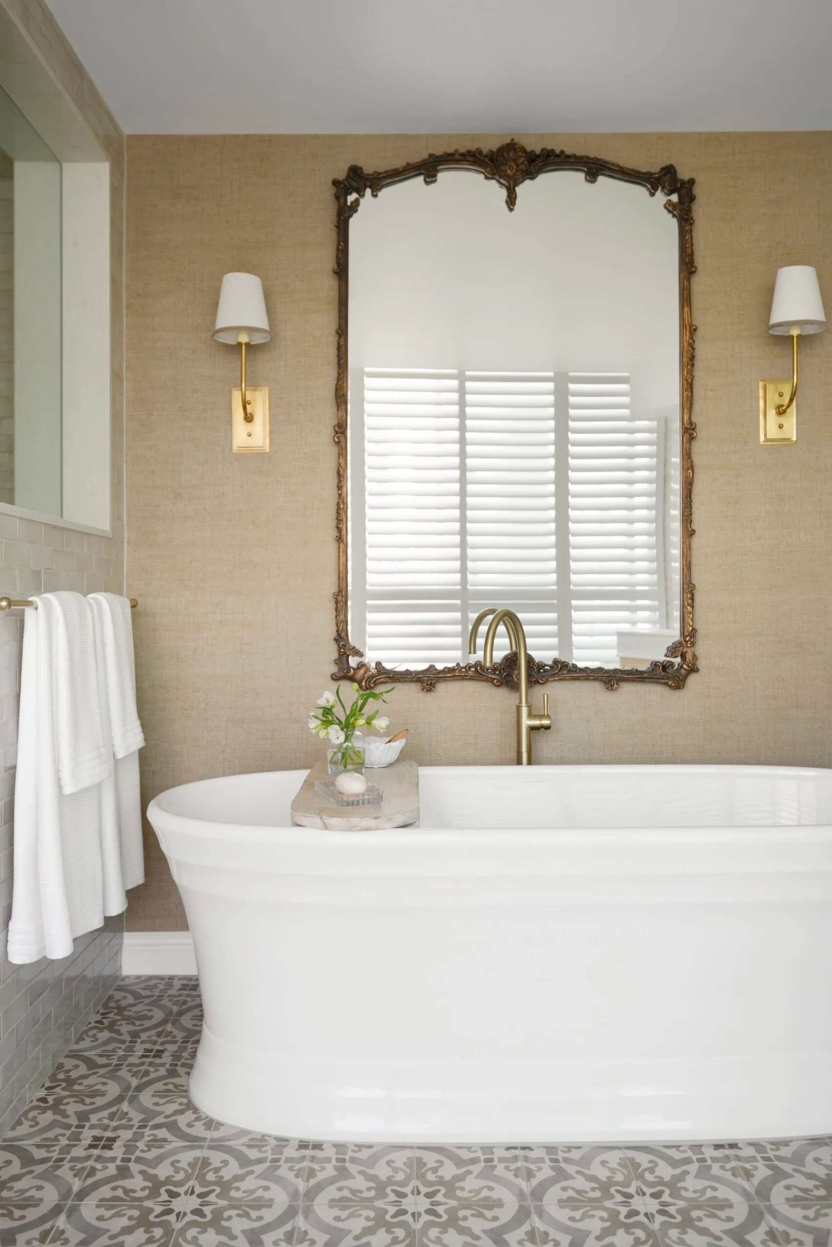 Rustic bathroom with white freestanding tub, ornate mirror, sconces, and patterned tile floor.