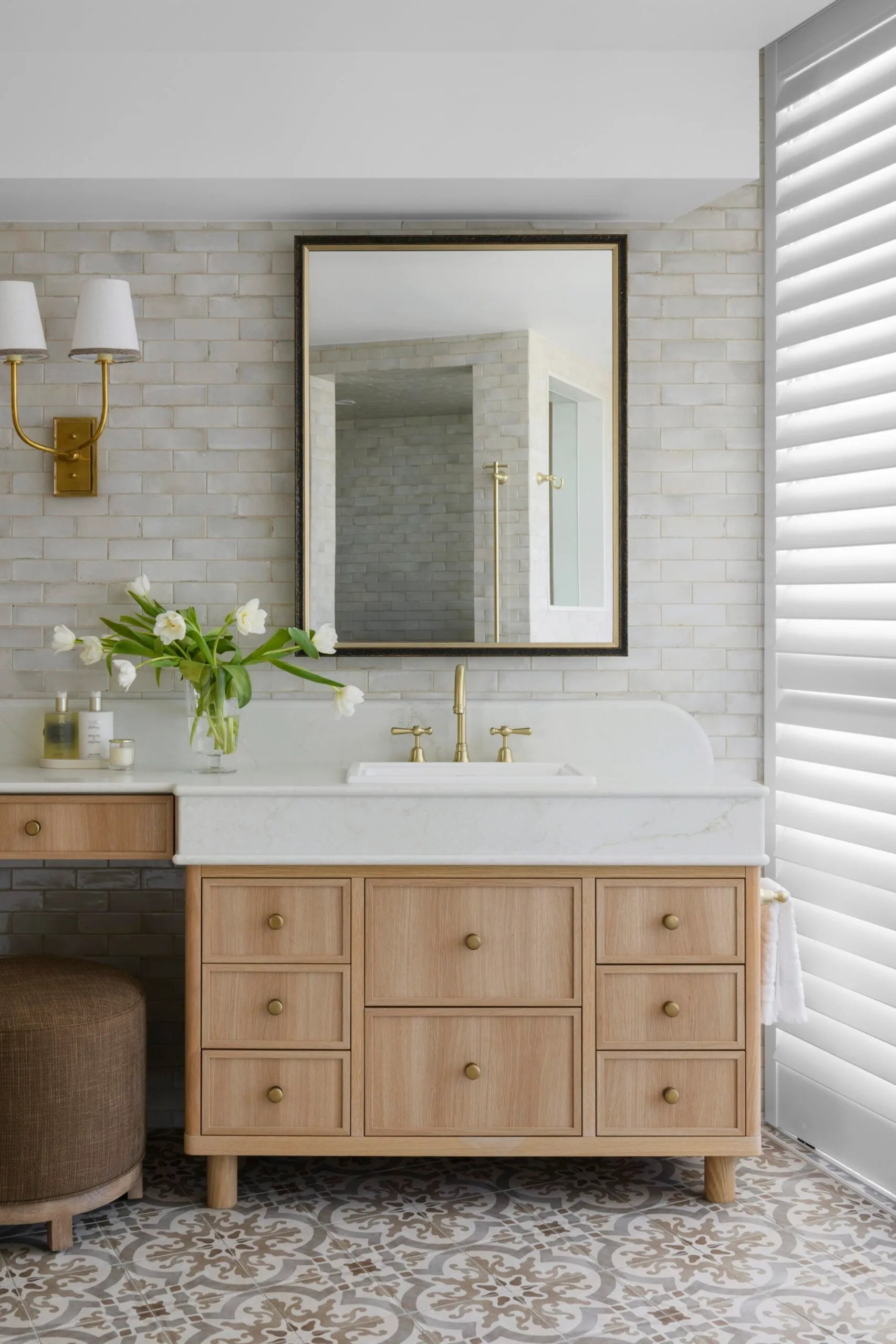 Stylish bathroom with a light wood vanity, brass fixtures, large mirror, and a vase of white tulips on a patterned tile floor.