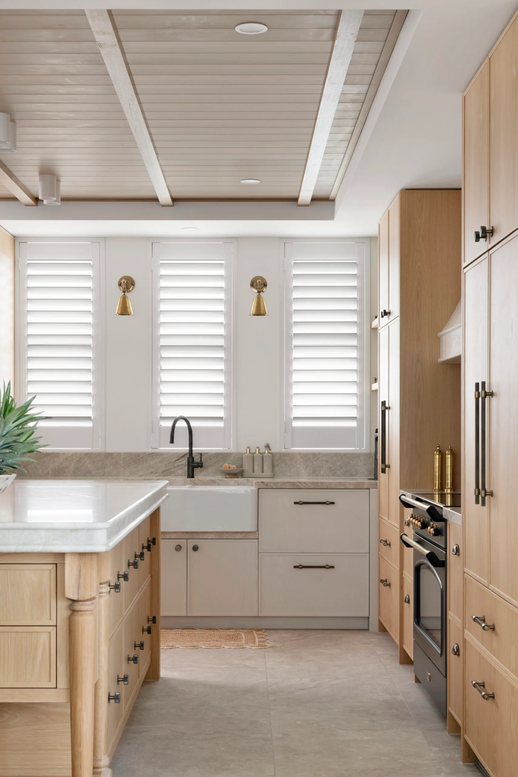 Modern kitchen interior with wooden cabinets, marble countertops, white shutters, gold sconces, and a black faucet.