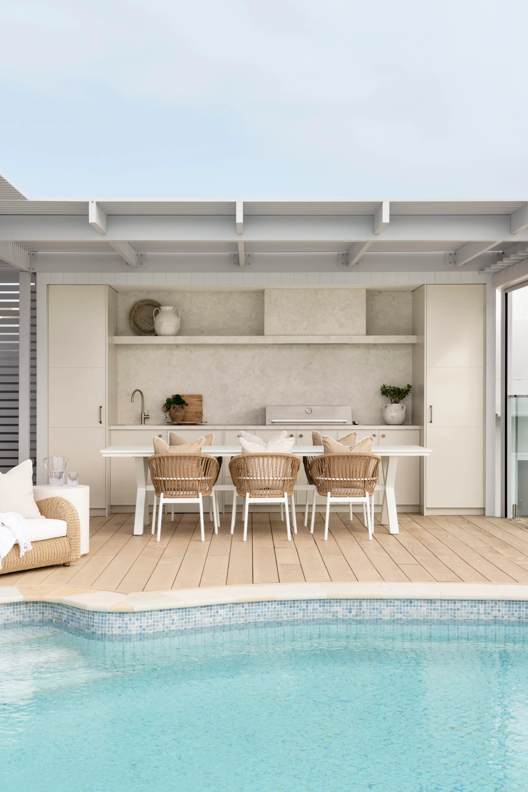 Outdoor kitchen with rattan chairs and a table on wooden decking by a clear blue pool.