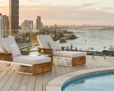 Sunset view from a rooftop pool deck with two lounge chairs, a wine bottle, glasses, and cityscape in the background. backyard pool ideas