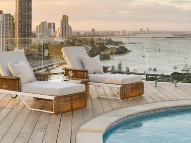 Sunset view from a rooftop pool deck with two lounge chairs, a wine bottle, glasses, and cityscape in the background. backyard pool ideas