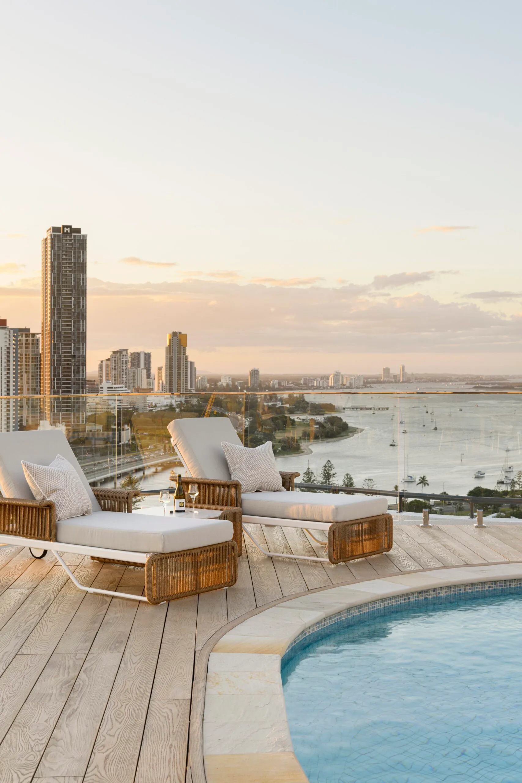 Sunset view from a rooftop pool deck with two lounge chairs, a wine bottle, glasses, and cityscape in the background.