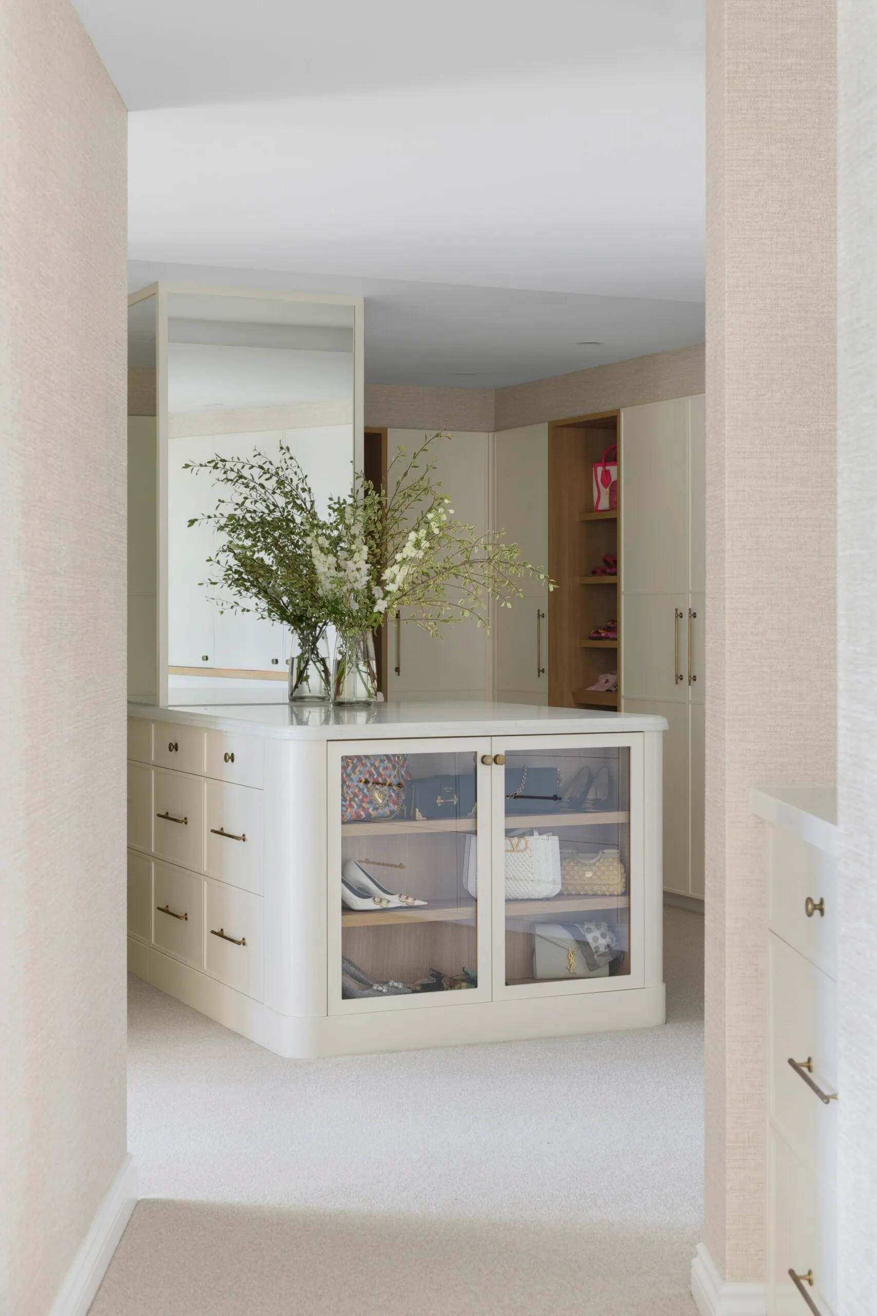 Luxurious walk-in closet with glass cabinet displaying shoes and bags, island counter with vase of flowers.