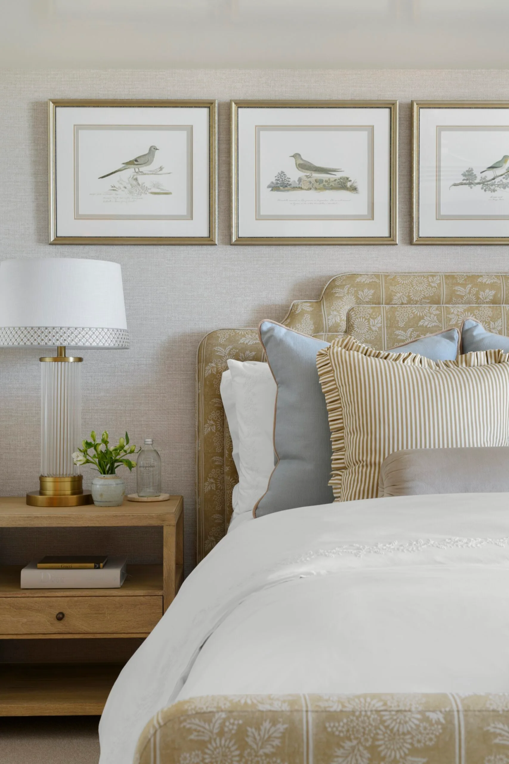 Bed with blue and striped pillows, wooden side table with lamp and plant. Bird artwork frames on wall.