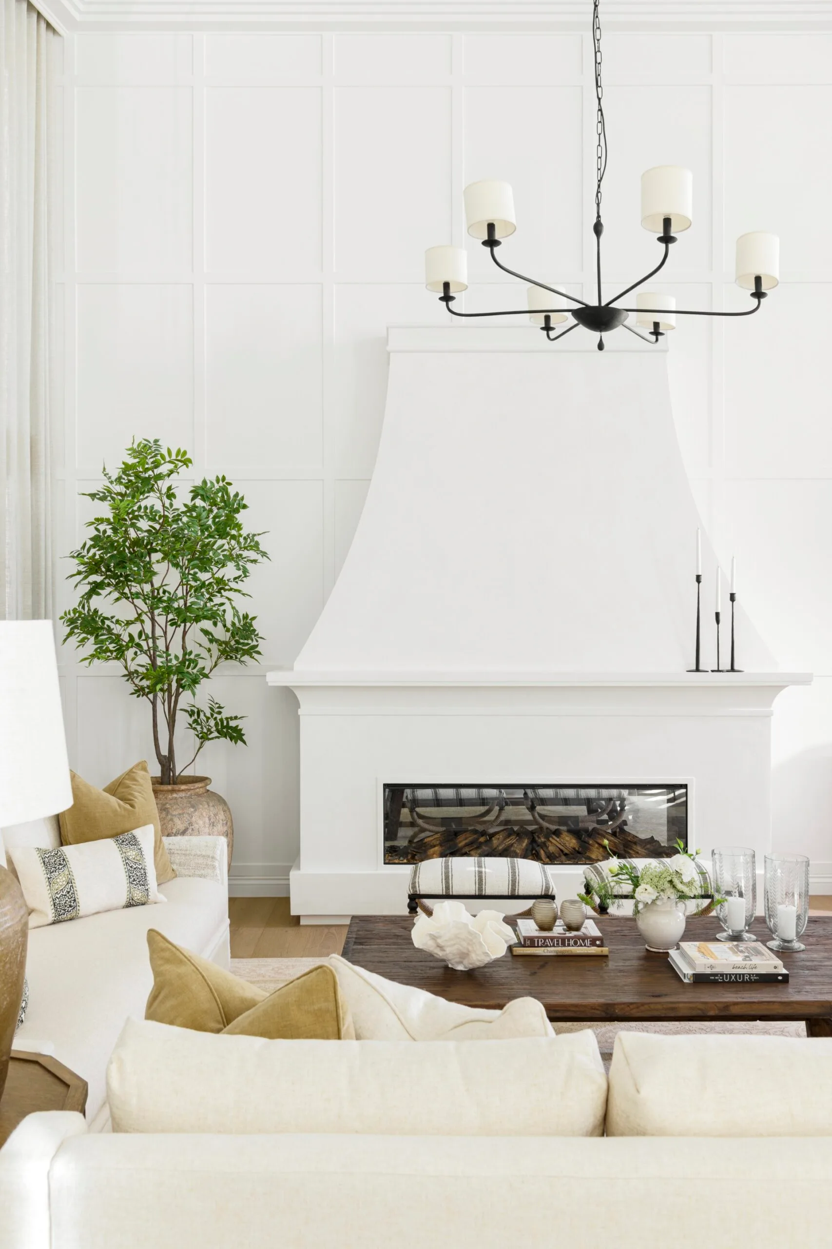 Bright living room with a white fireplace, potted plant, beige sofas, wooden coffee table, and black chandelier.