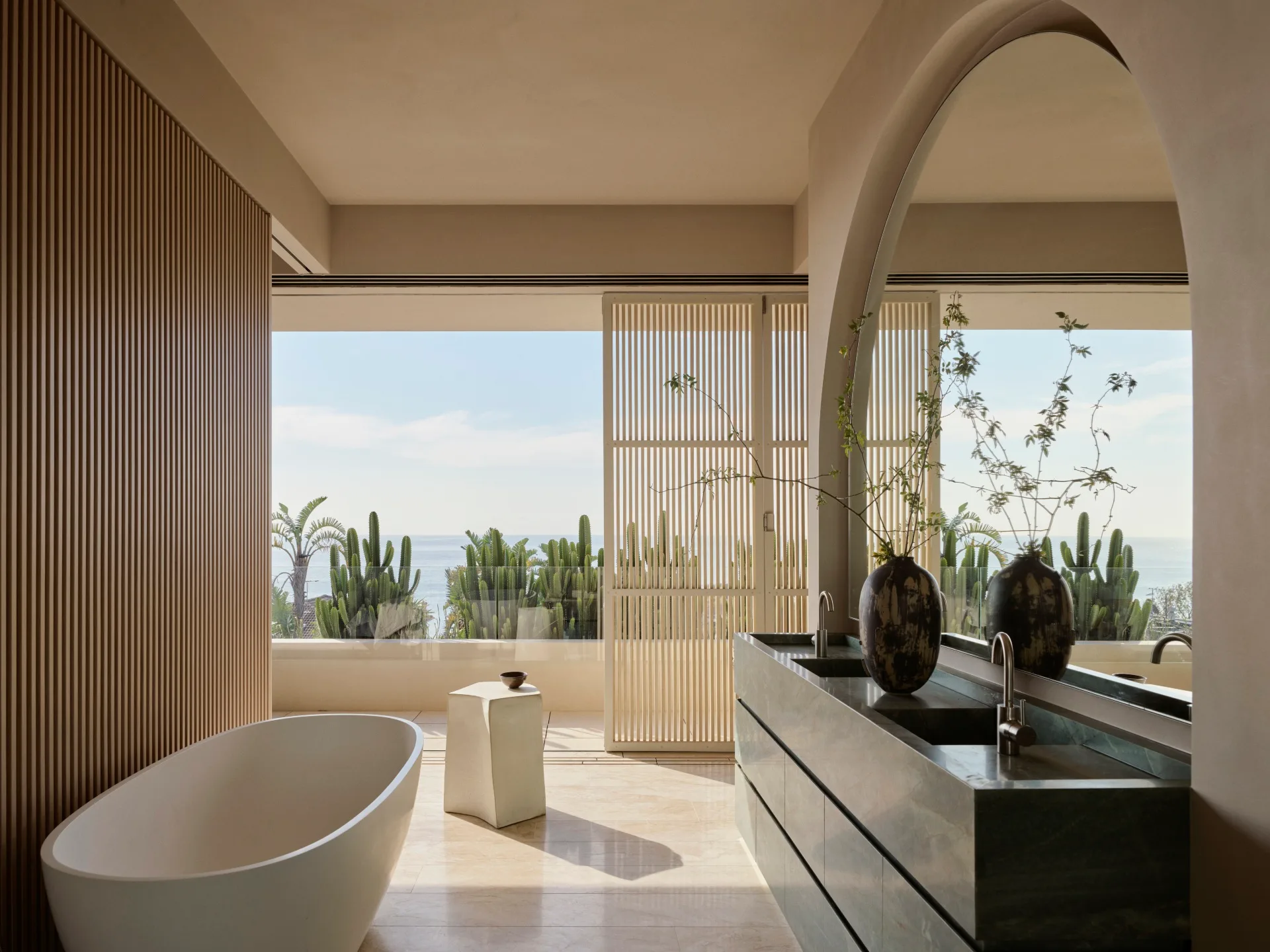 Luxurious bathroom with a large window view of the ocean, featuring a freestanding tub, modern sink, and cacti outside.