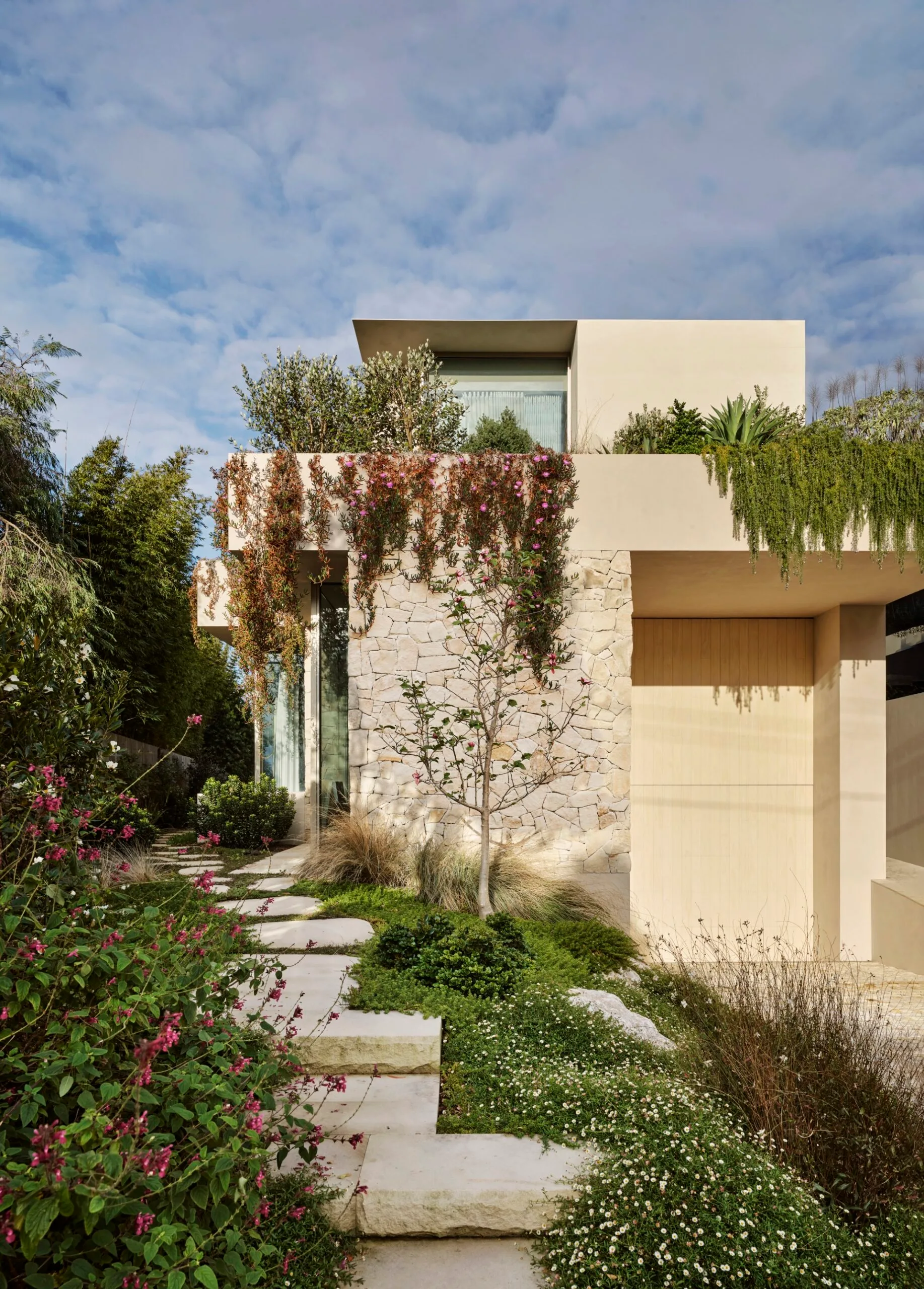 Modern two-story house with stone facade, lush garden, and flowering plants under a partly cloudy sky.