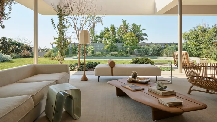 Spacious open-plan living room with modern furniture, large windows, and garden view, blending indoor and outdoor spaces. A wooden coffee table is in the middle of the room, with two white sofas nearby. The backyard is visible, with a pool in the background.