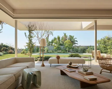 Spacious open-plan living room with modern furniture, large windows, and garden view, blending indoor and outdoor spaces. A wooden coffee table is in the middle of the room, with two white sofas nearby. The backyard is visible, with a pool in the background.