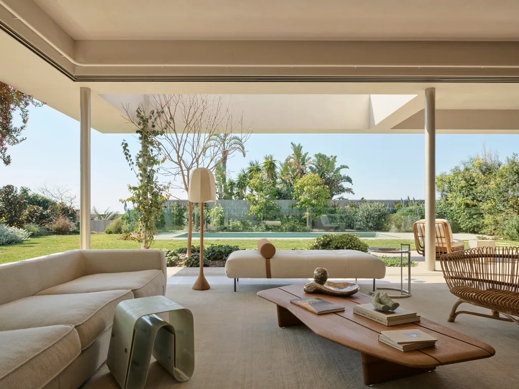 Spacious open-plan living room with modern furniture, large windows, and garden view, blending indoor and outdoor spaces. A wooden coffee table is in the middle of the room, with two white sofas nearby. The backyard is visible, with a pool in the background.