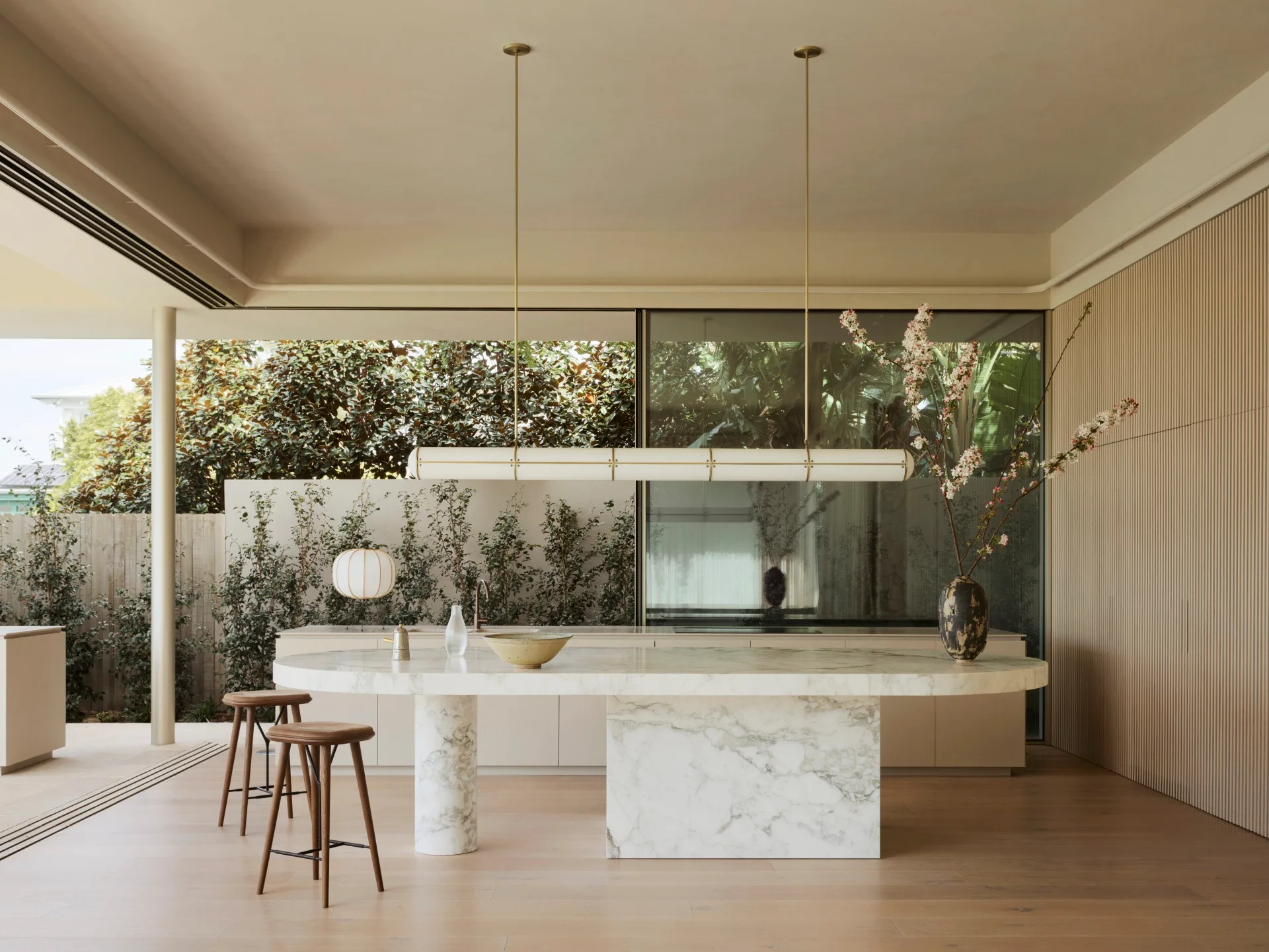Modern open-plan kitchen with a marble island, bar stools, pendant lighting, and greenery visible through large glass doors that slide open for indoor-outdoor flow.