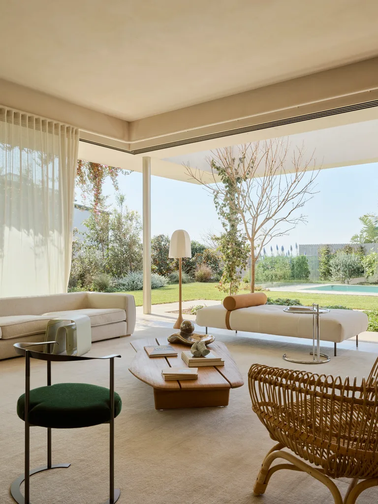 Contemporary open-plan living room with modern furniture, a large window view of a garden, and a pool beyond. A wooden coffee table is in the middle of the room, with two white sofas nearby.