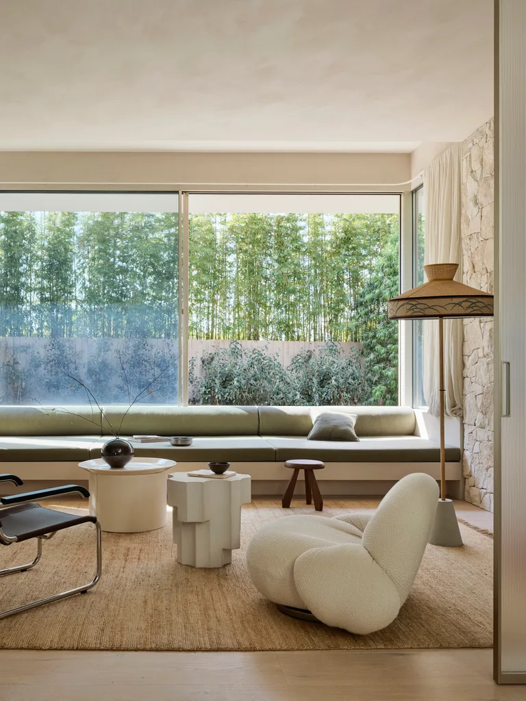 Modern living room with a sculptural white armchair, two coffee tables, large window view, greenery outside, and earthy decor. A sisal rug is on the wooden floorboards.