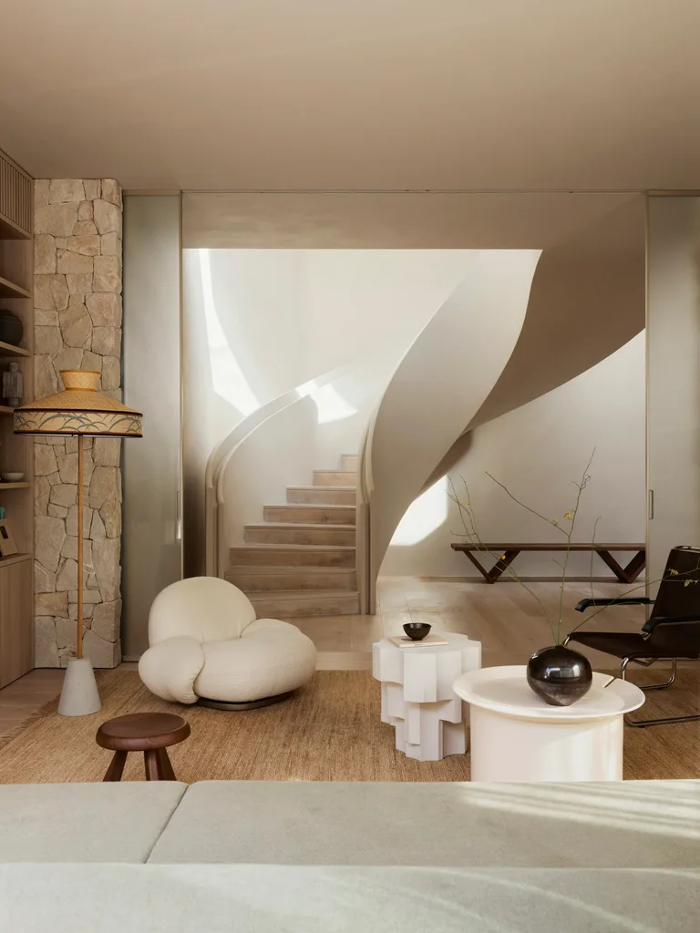 Open-plan living area with a curved staircase, plush white armchair, and decorative tables on a neutral-toned rug.