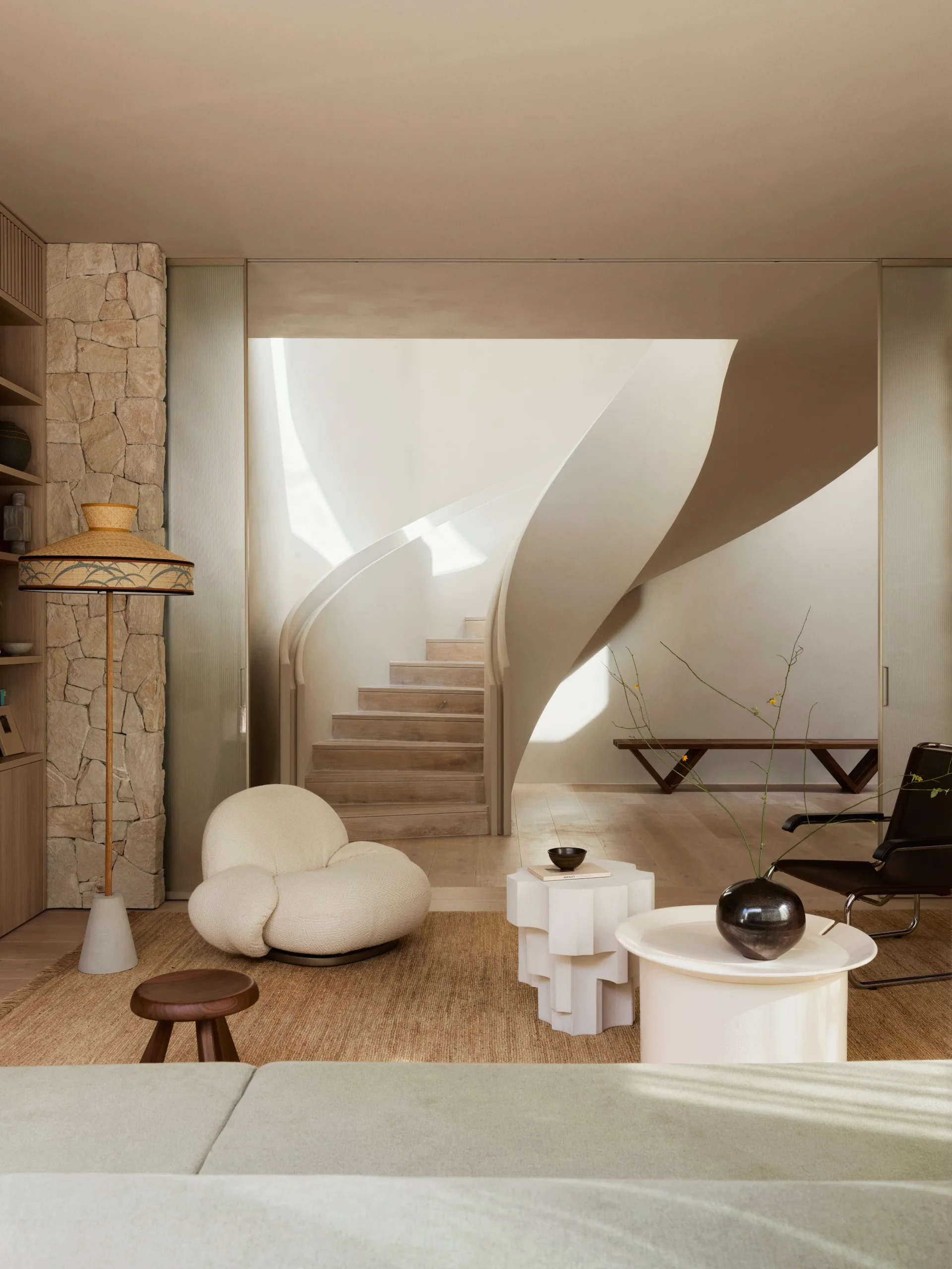Open-plan living area with a curved staircase, plush white armchair, and decorative tables on a neutral-toned rug.