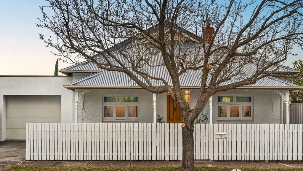 White single-story house with a metal roof, fronted by a tree and picket fence.