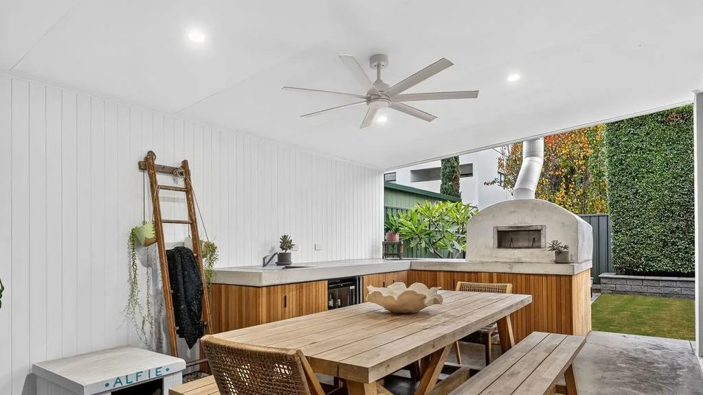 Outdoor patio with wooden dining set, ceiling fan, pizza oven, lush greenery, and a ladder-style plant shelf.