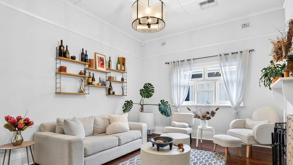 Living room with white sofa, armchairs, wooden shelves, plants, and large window with sheer curtains.