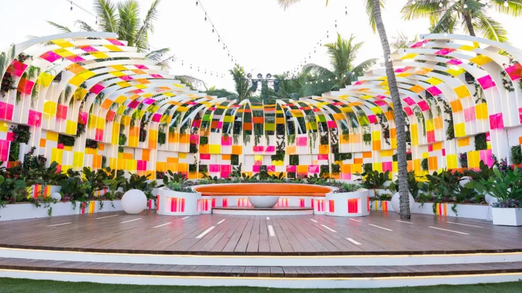 Colorful outdoor firepit area in the Love Island USA villa with vibrant panels, tropical plants, and an orange semicircular bench. Palm trees in the background.