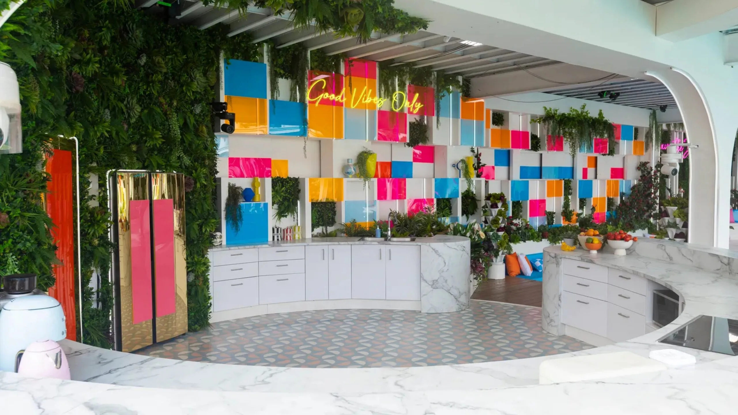Colorful kitchen in the 2026 Love Island USA villa with greenery, vibrant wall panels, marble countertops, and "Good Vibes Only" neon sign.