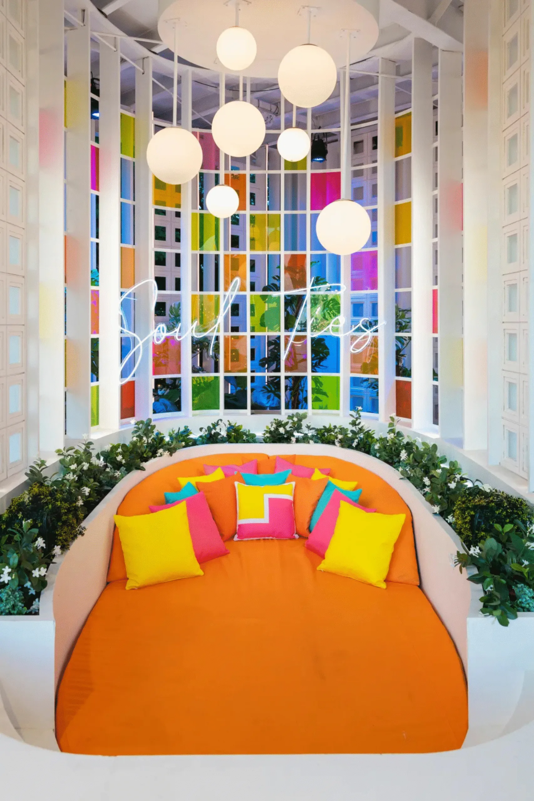 A bright and bold daybed, with orange bedding and a mix of yellow, pink and blue cushions. Fake plants surround the white daybed platform and round light pendants are above it. A neon signs says 'soul ties'. This is part of the Love Island USA villa in 2026.