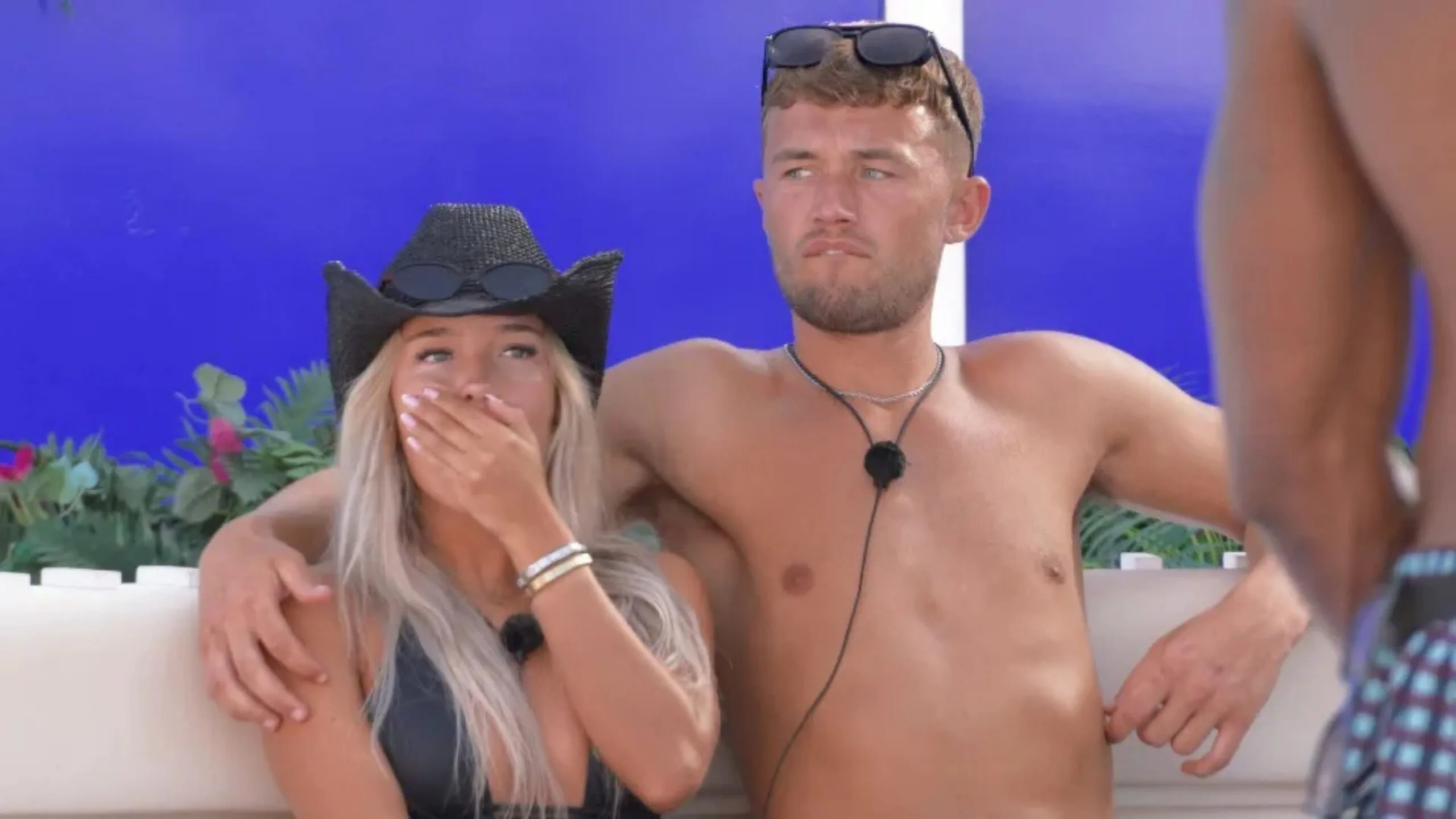 A shirtless man and woman in a hat sit on a couch outside; the woman covers her mouth with her hand. This is a screengrab from Love Island.