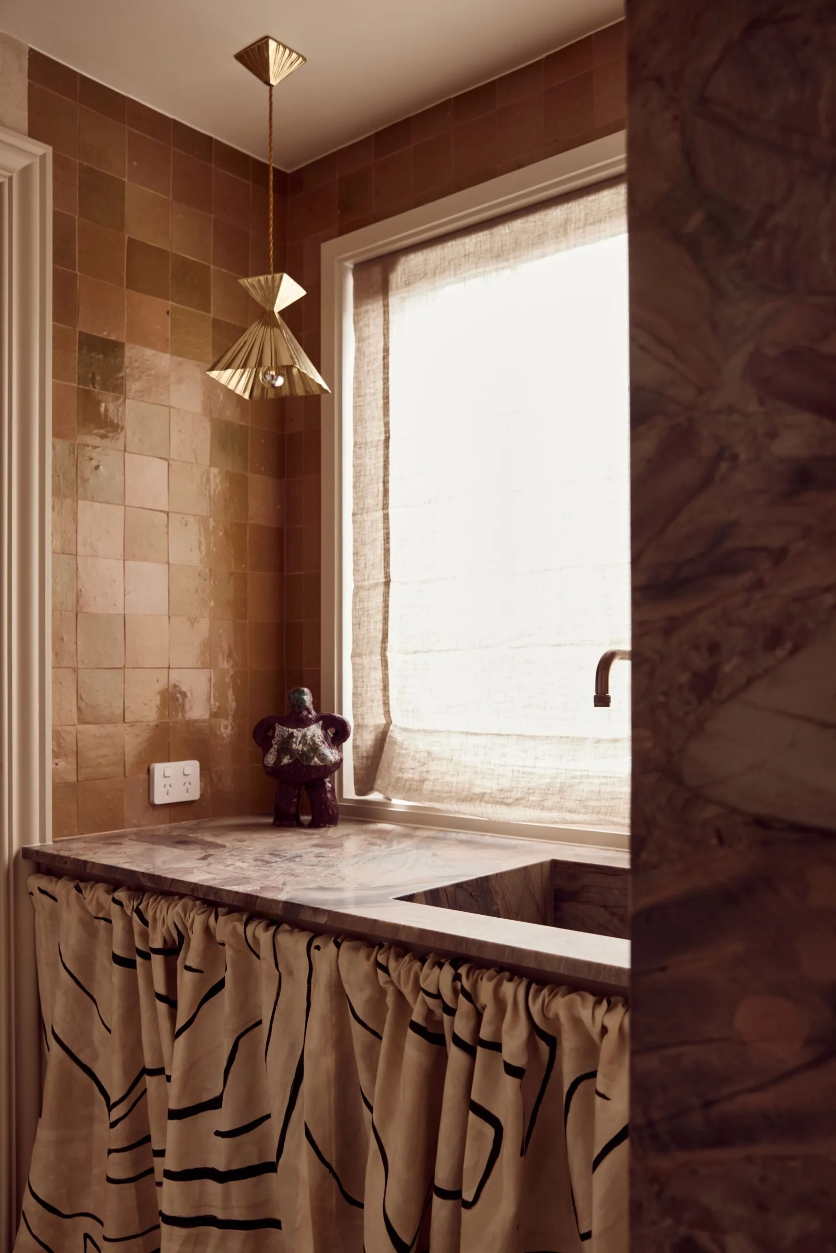 A brown kitchen with a sink skirt, brown tiles and a gold pendant light