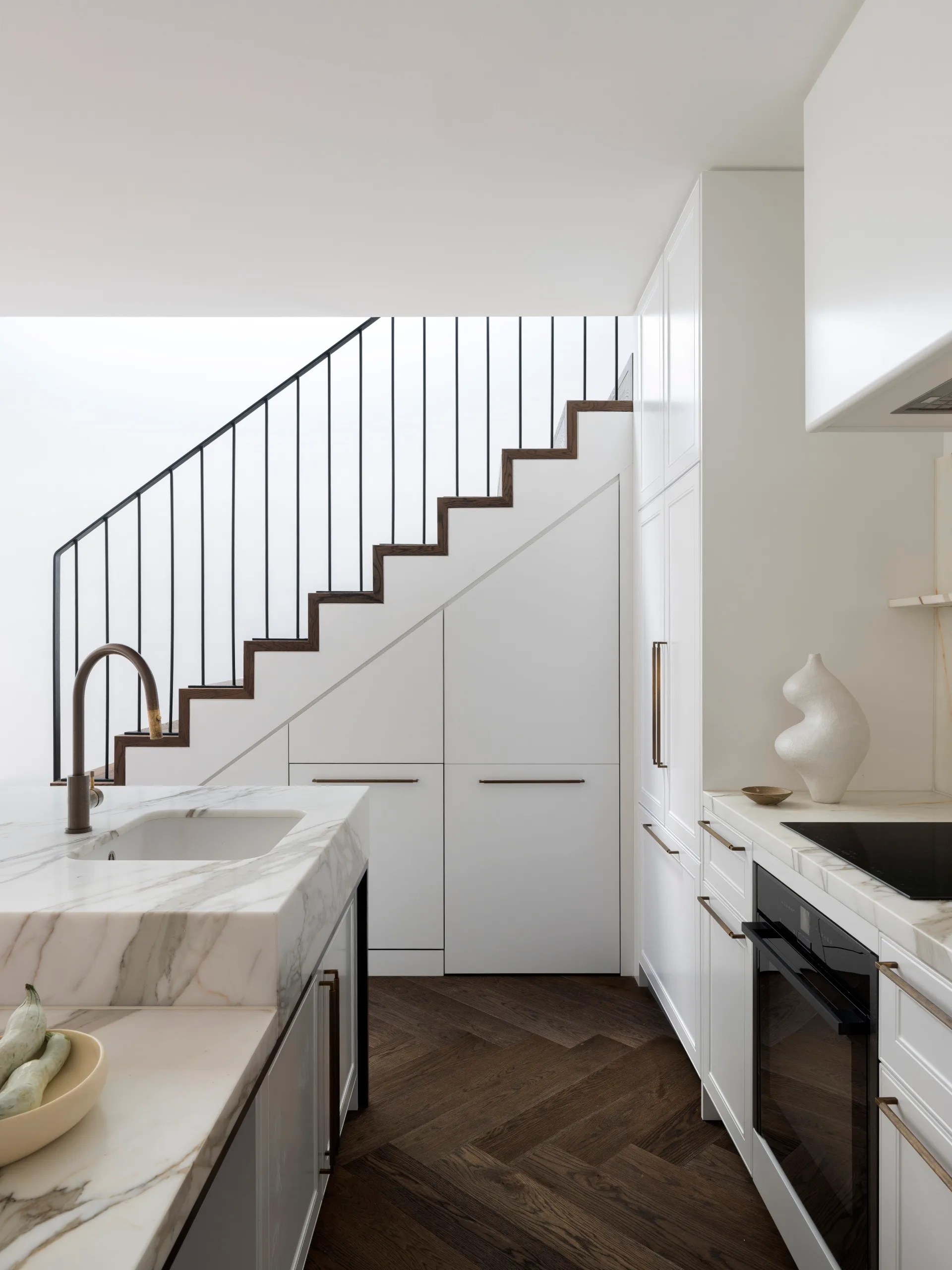 A modern, minimalist kitchen with a dual-height island, marble benchtops, under-stair storage, and dark herringbone flooring.