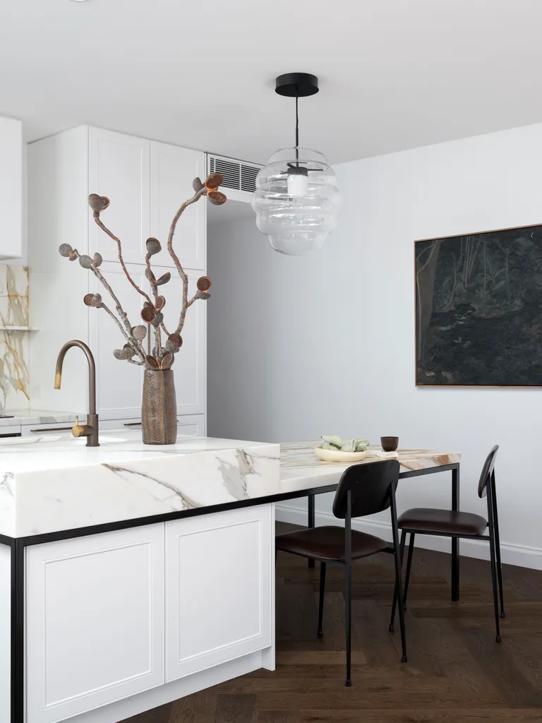 Modern kitchen with white cabinets, a double-height marble benchtop, pendant light, vase with branches, artwork and two dining chairs.
