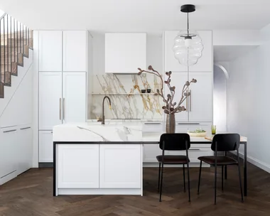 A modern kitchen with white cabinets, a marble island, dark wood flooring, pendant light, and two chairs.