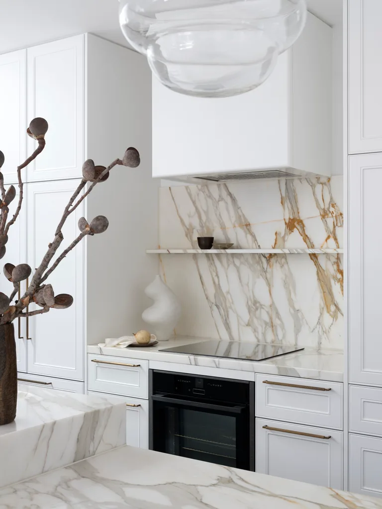 A modern kitchen with white joinery, Calacatta Gold marble benchtops and splashback, a vase with branches, and a sleek black oven.
