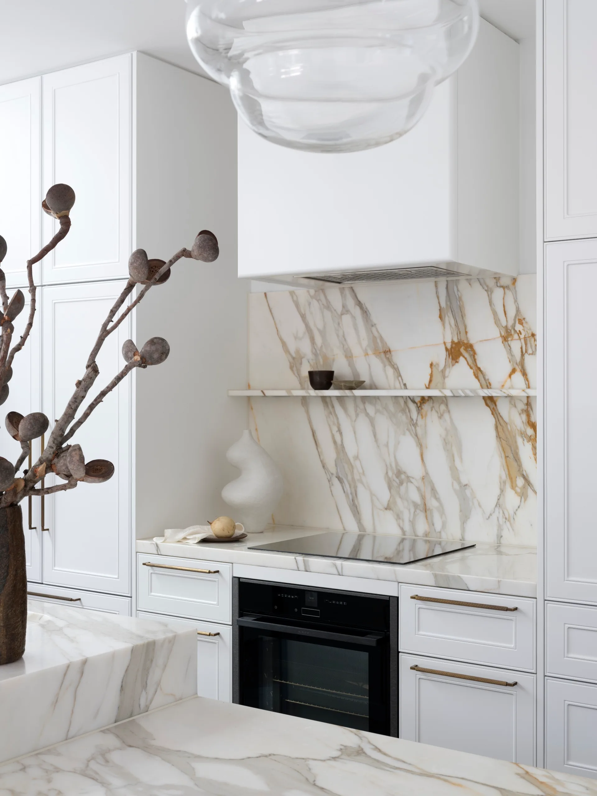 A modern kitchen with white joinery, Calacatta Gold marble benchtops and splashback, a vase with branches, and a sleek black oven.