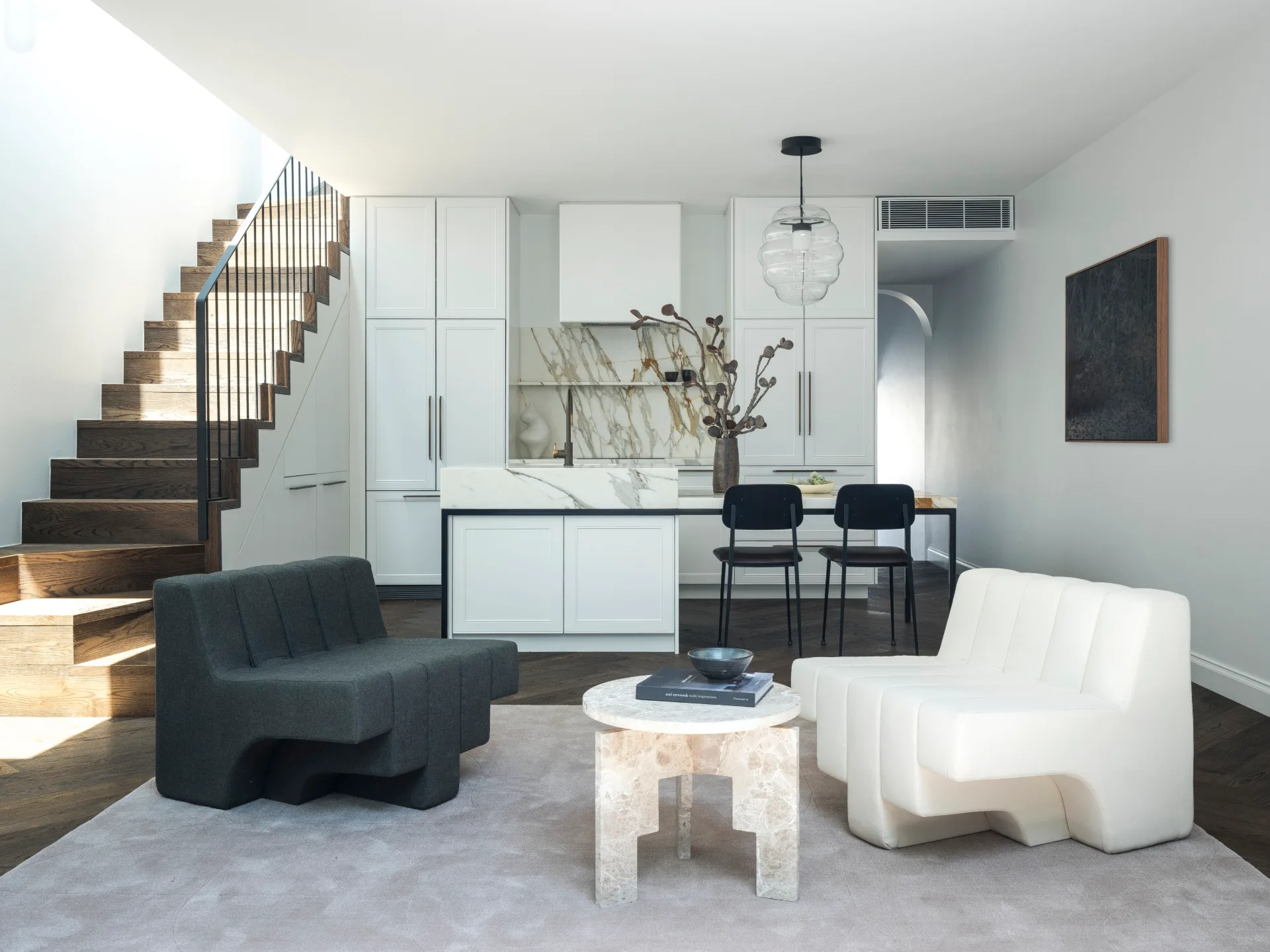 Modern open-plan living area with dark timber stairs, white kitchen cabinets, black and white statement armchairs, and a marble coffee table.