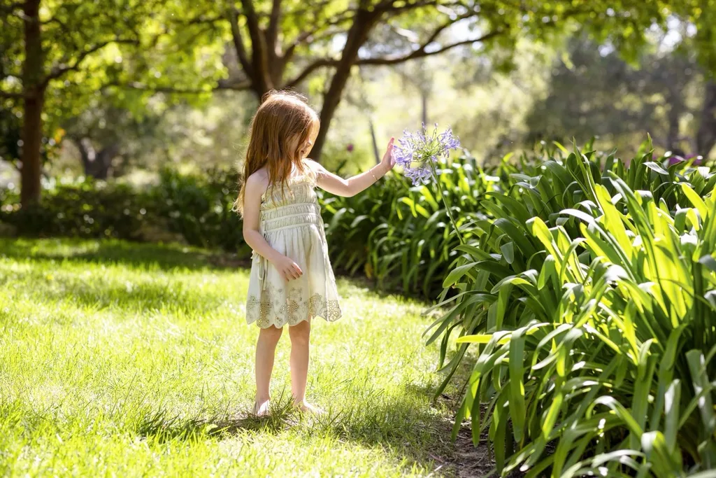 Princess Lilibet stands barefoot in a lush garden beside flowering purple agapanthus.