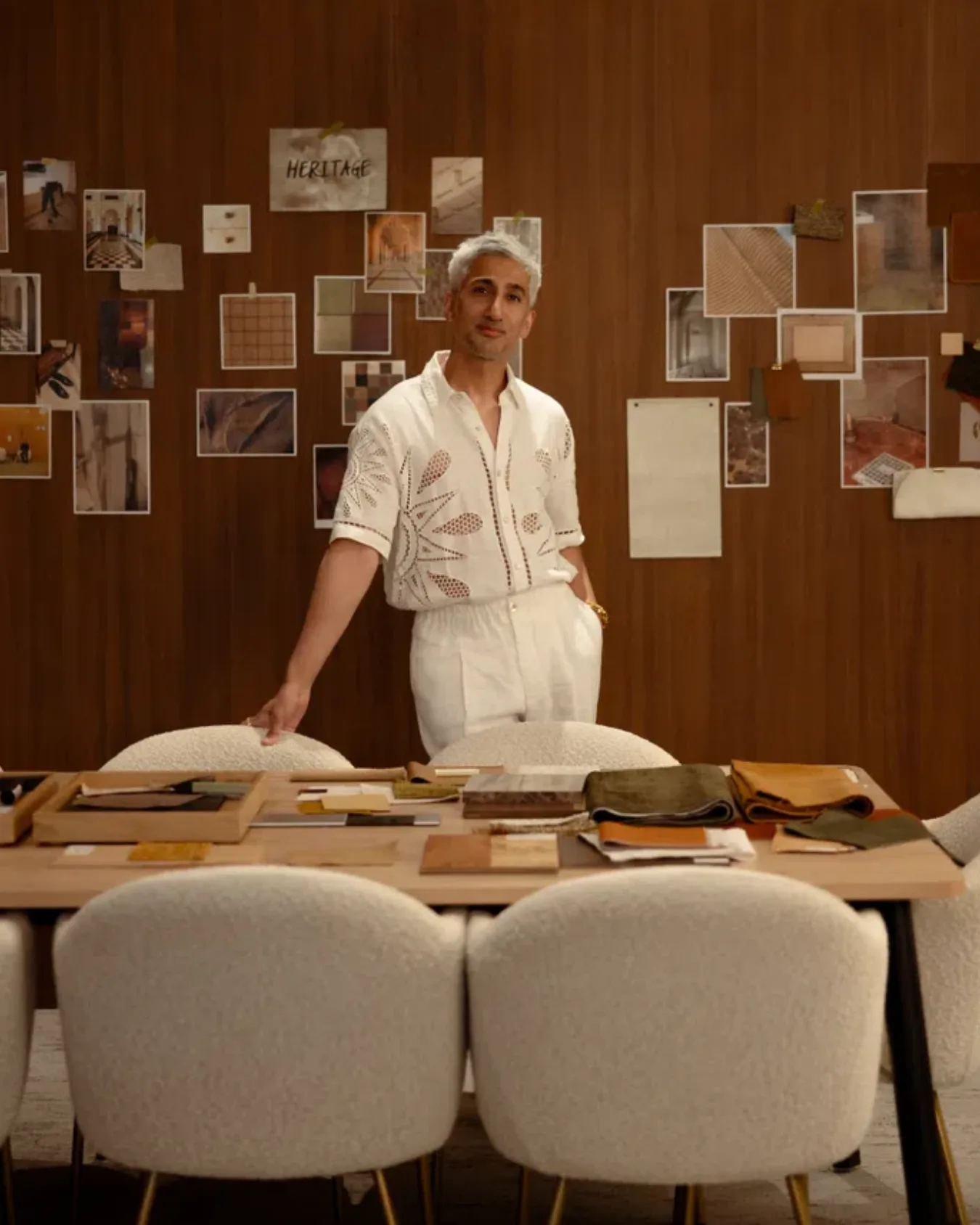 Tan france stands behind a light wood desk surrounded by chairs. On the back wall, a collection of fabrics and images are cellotaped to the wall
