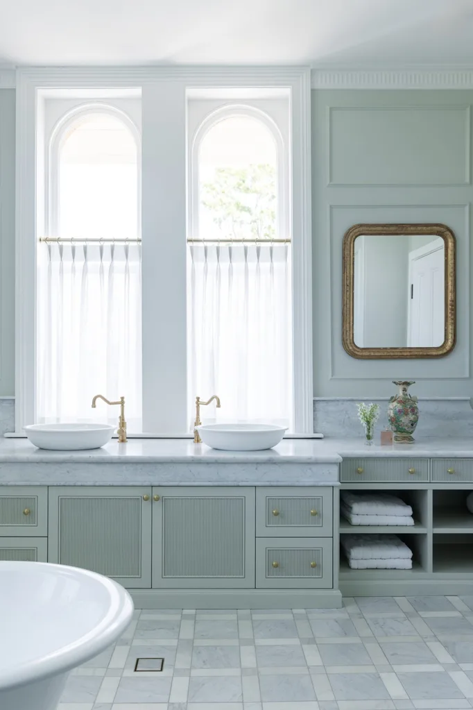 Elegant bathroom with double sinks, marble countertop, gold faucets, large arched windows with sheer cafe curtains, gold framed mirror, and decorative vase.