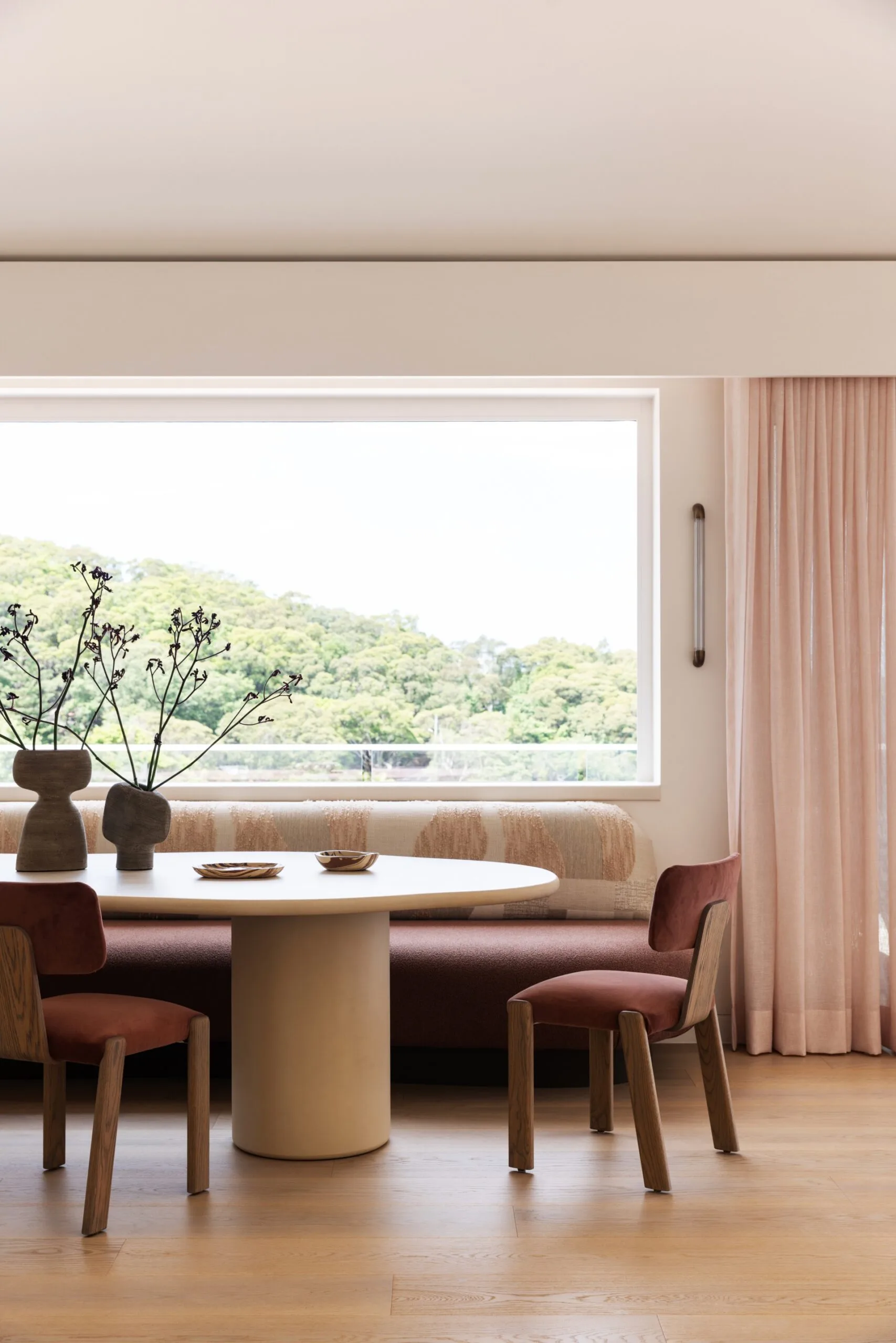 Modern breakfast nook with a beautiful-in upholstered banquette, a round table, wooden chairs, and decorative vases. Large window with a nature view positioned above, with sheer pink curtains to the side.