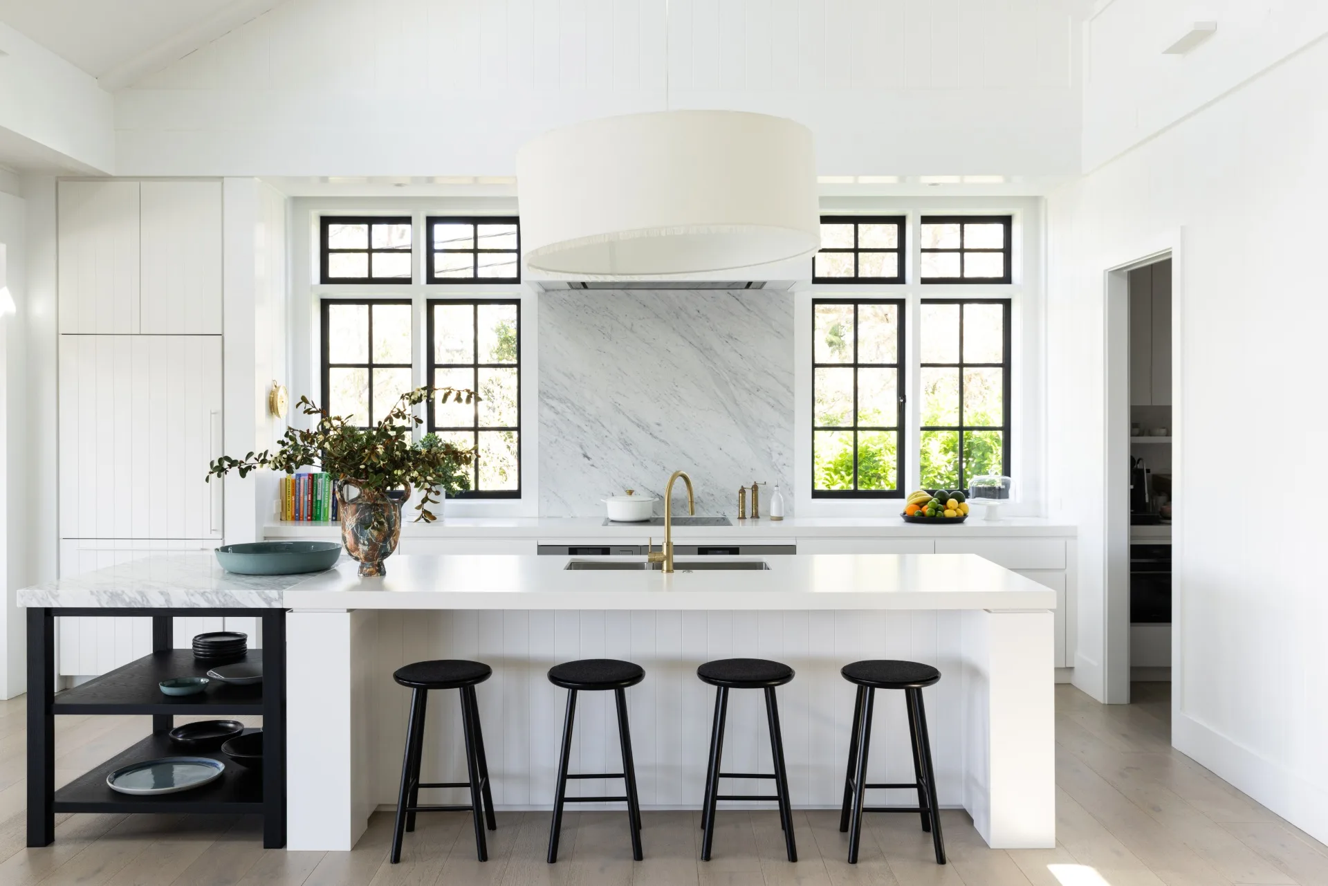 Modern white kitchen with a large island, black stools, large window, and decorative vase on the counter.