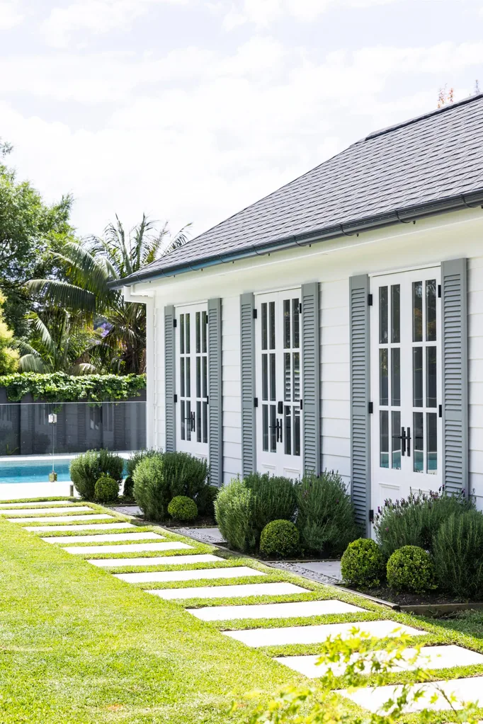 Modern house with white exterior, gray shutters, large windows, garden, and pool in background under a blue sky.
