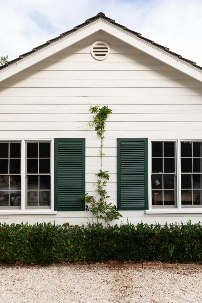 White house exterior with green shutters and climbing vine, set against a gravel pathway and trimmed hedges.
