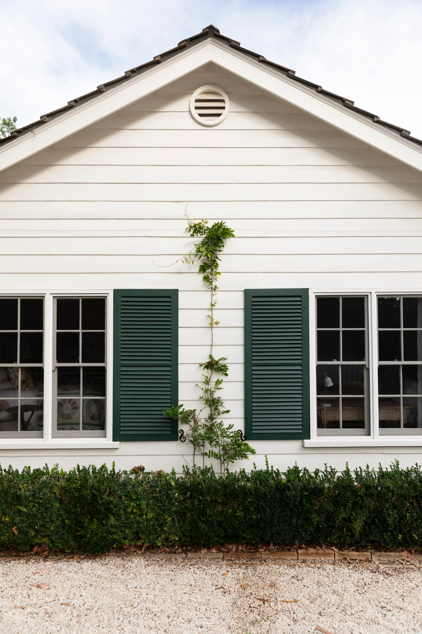 White house exterior with green shutters and climbing vine, set against a gravel pathway and trimmed hedges.
