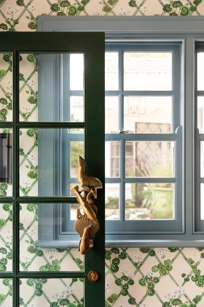 Glass door with green timber frame and panes with brass sea-themed handle, patterned wallpaper, and blue-framed window and panes revealing a garden view.