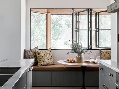 Cozy kitchen nook with built-in banquette seating, patterned cushions, a round table, flowers, and open windows. Blue joinery in a galley shaped kitchen is just visible in the foreground.